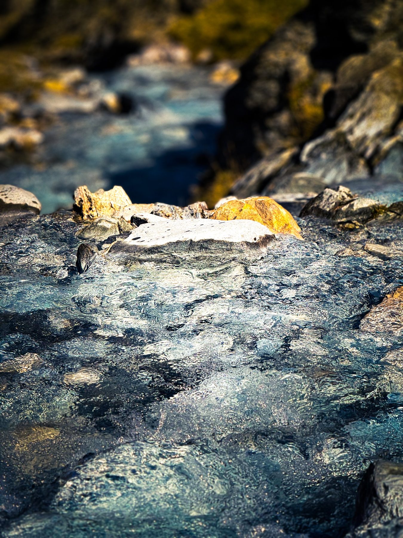 A ras — arroyo de montaña fotografiado a ras del agua, con la corriente azul y transparente corriendo sobre las piedras y unas rocas doradas por el sol al fondo desenfocado, en color