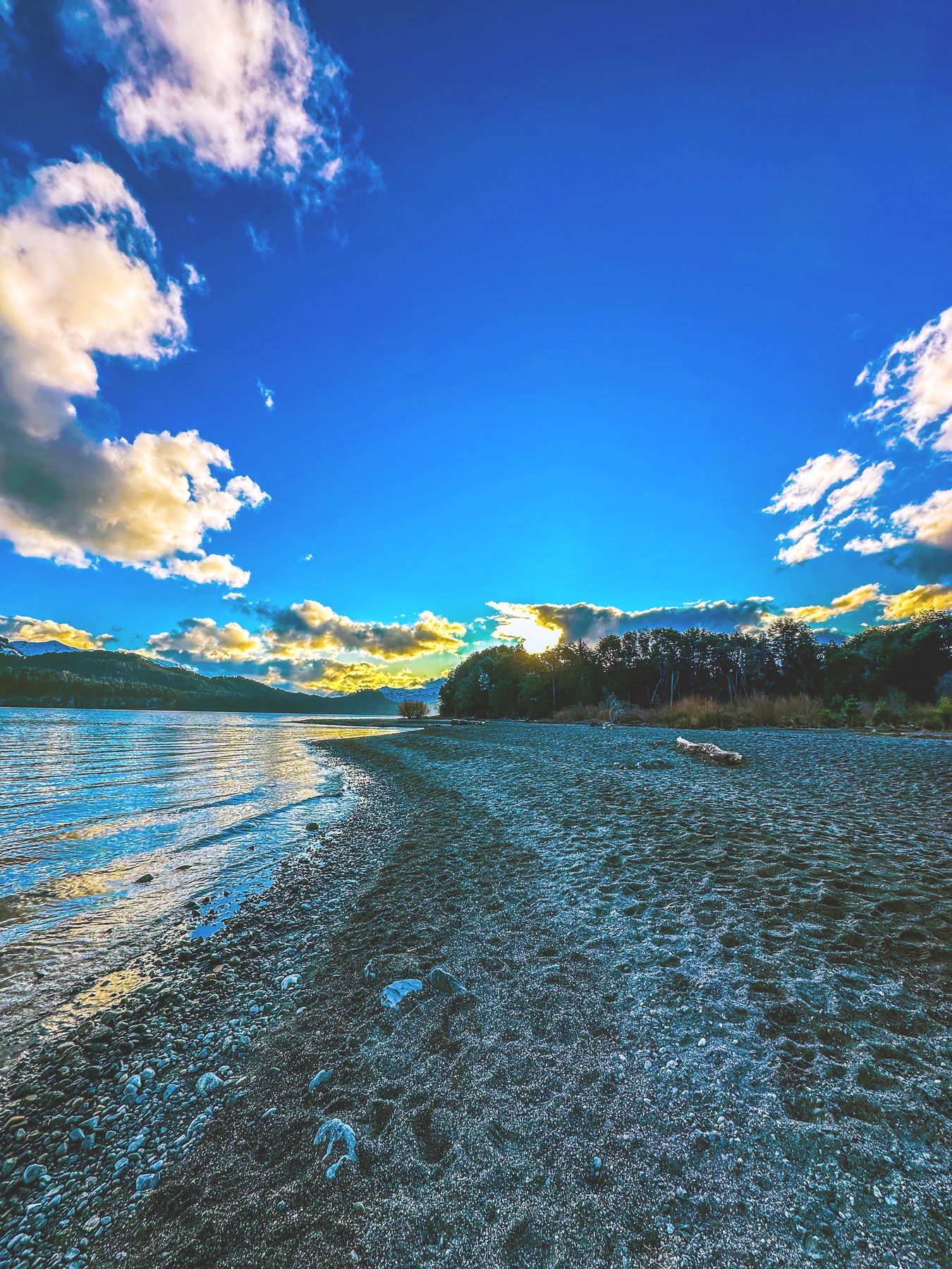 Atardecer en el lago Nahuel Huapi — orilla de arena y agua quieta bajo un cielo azul intenso con nubes, y la luz del sol bajo sobre la cordillera, en color