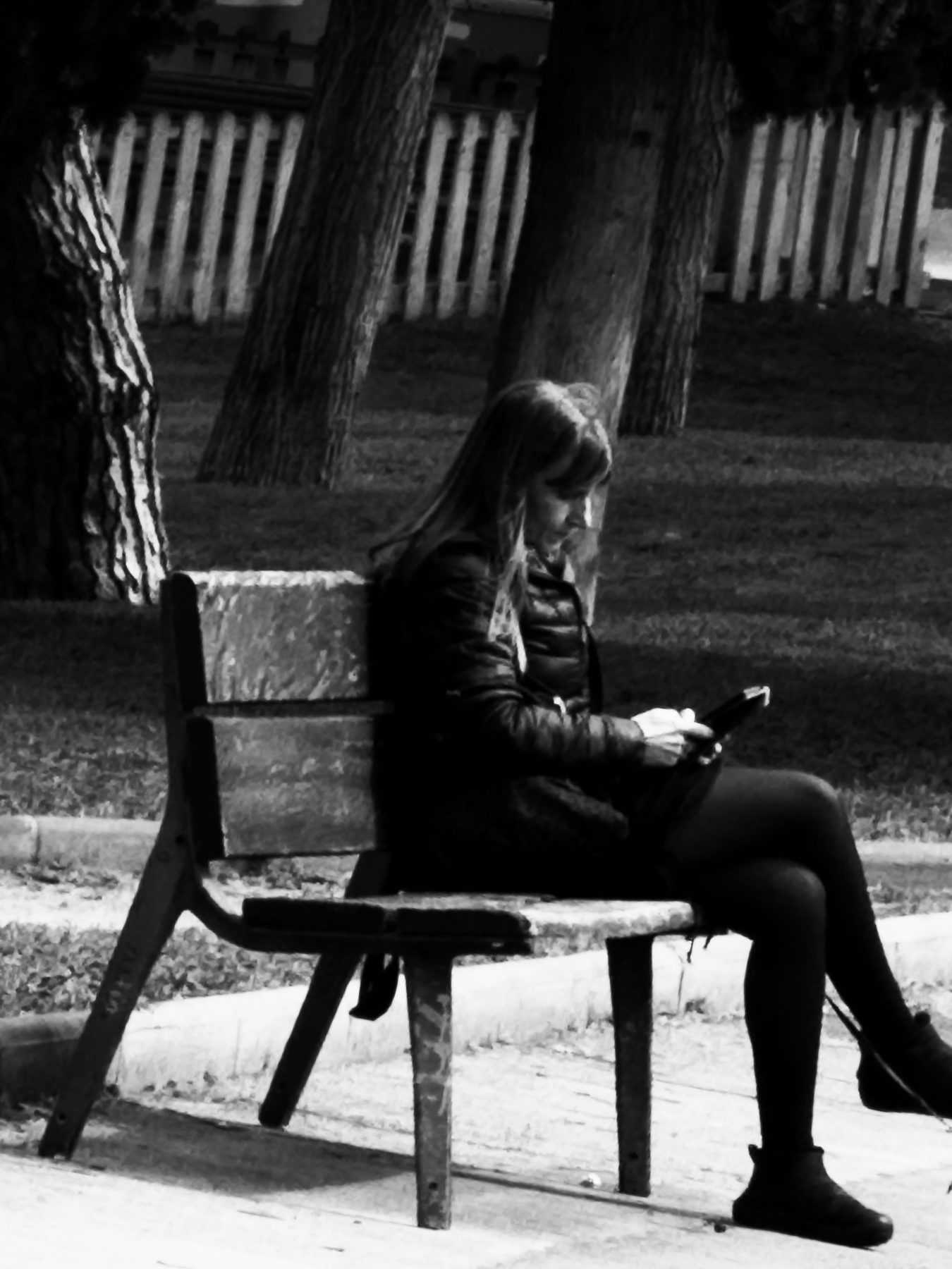A la sombra — mujer sentada de perfil en un banco de parque mirando el móvil, entre troncos de árbol, en blanco y negro