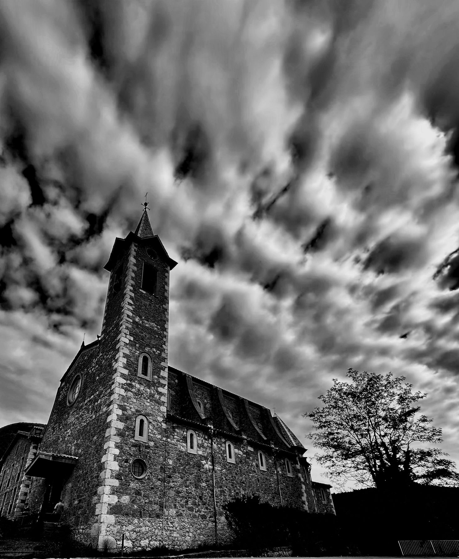 Campanario — iglesia de piedra con torre puntiaguda bajo un cielo de nubes rasgadas, en blanco y negro