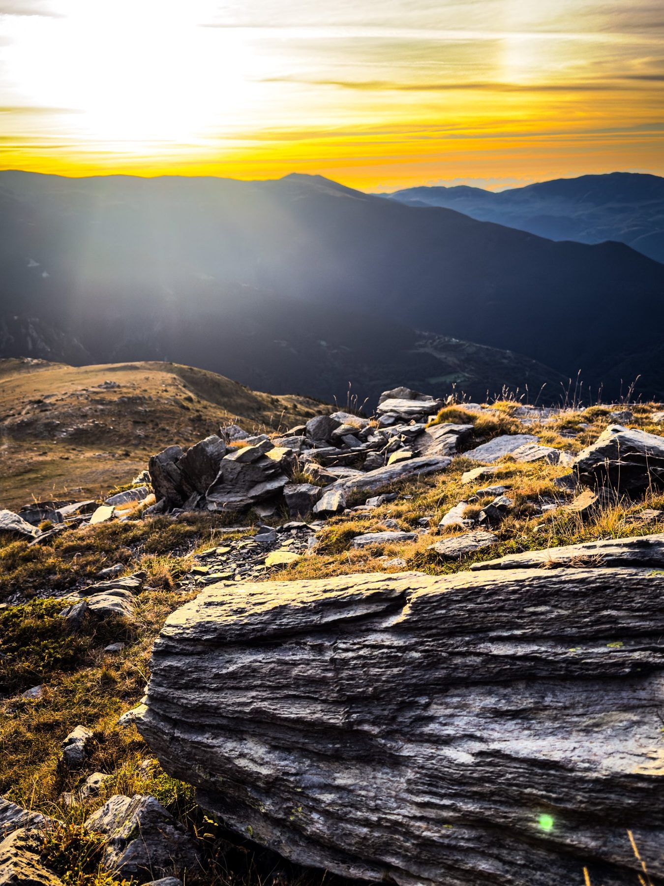 Capas — roca de pizarra en capas en primer plano sobre una cumbre con hierba seca, y detrás varias filas de montañas en sombra bajo un cielo amarillo con el sol entre nubes, en color