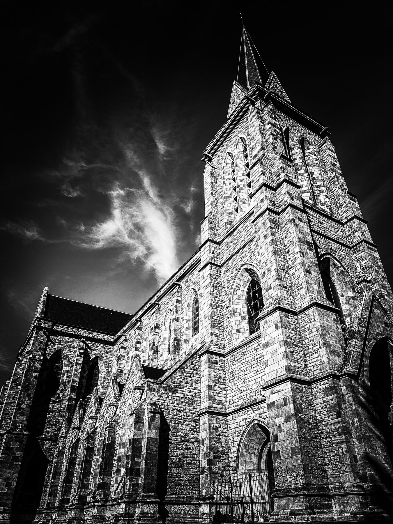 Catedral de Bariloche — torre y nave de piedra vistas desde abajo, con el campanario apuntando a un cielo oscuro cruzado por una nube blanca, en blanco y negro