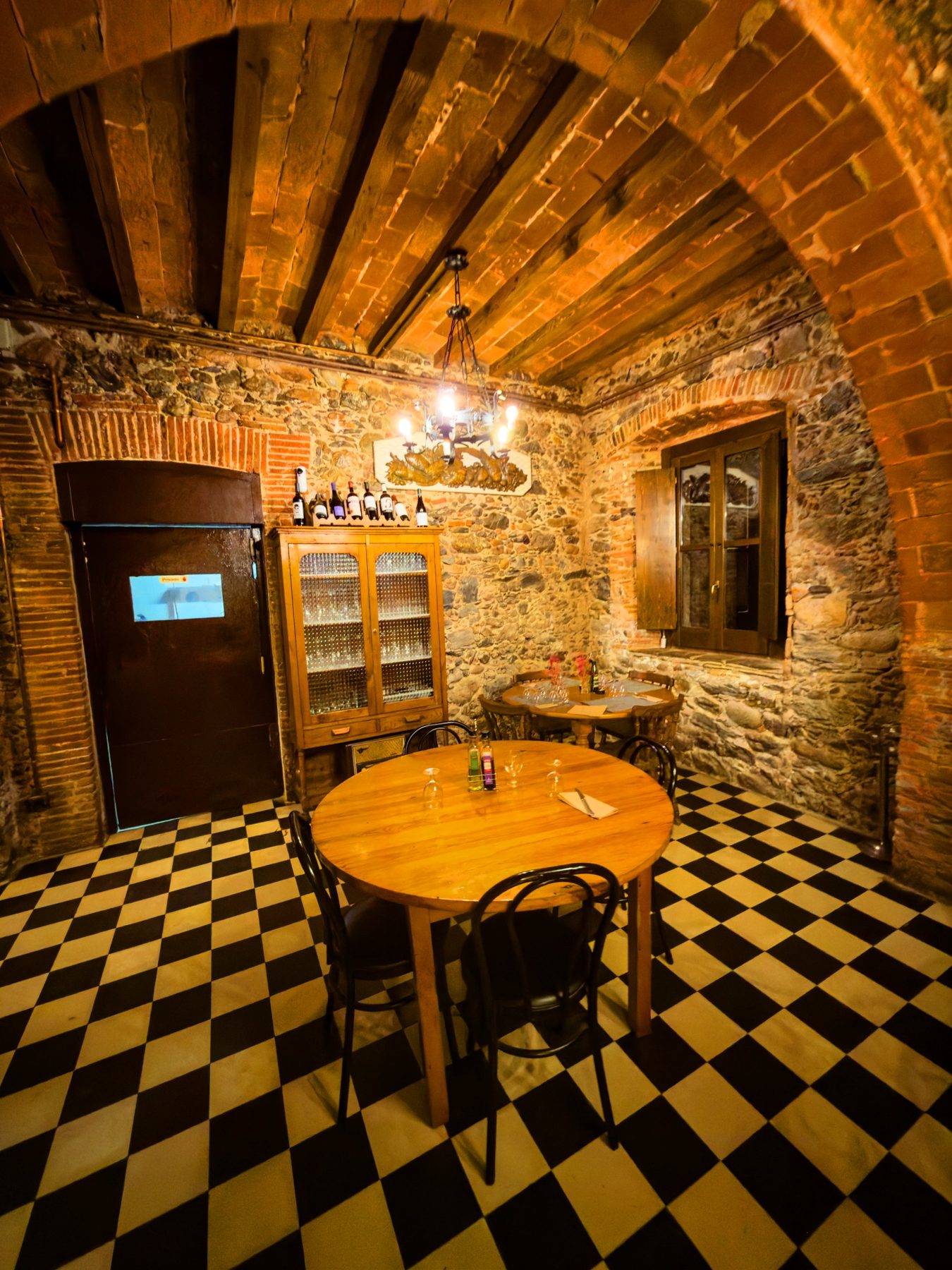 Lustros de sobremesa — salón antiguo de un restaurante de montaña visto a través de un arco de ladrillo, con techo de vigas, paredes de piedra, suelo de damero blanco y negro y mesas de madera puestas, en color