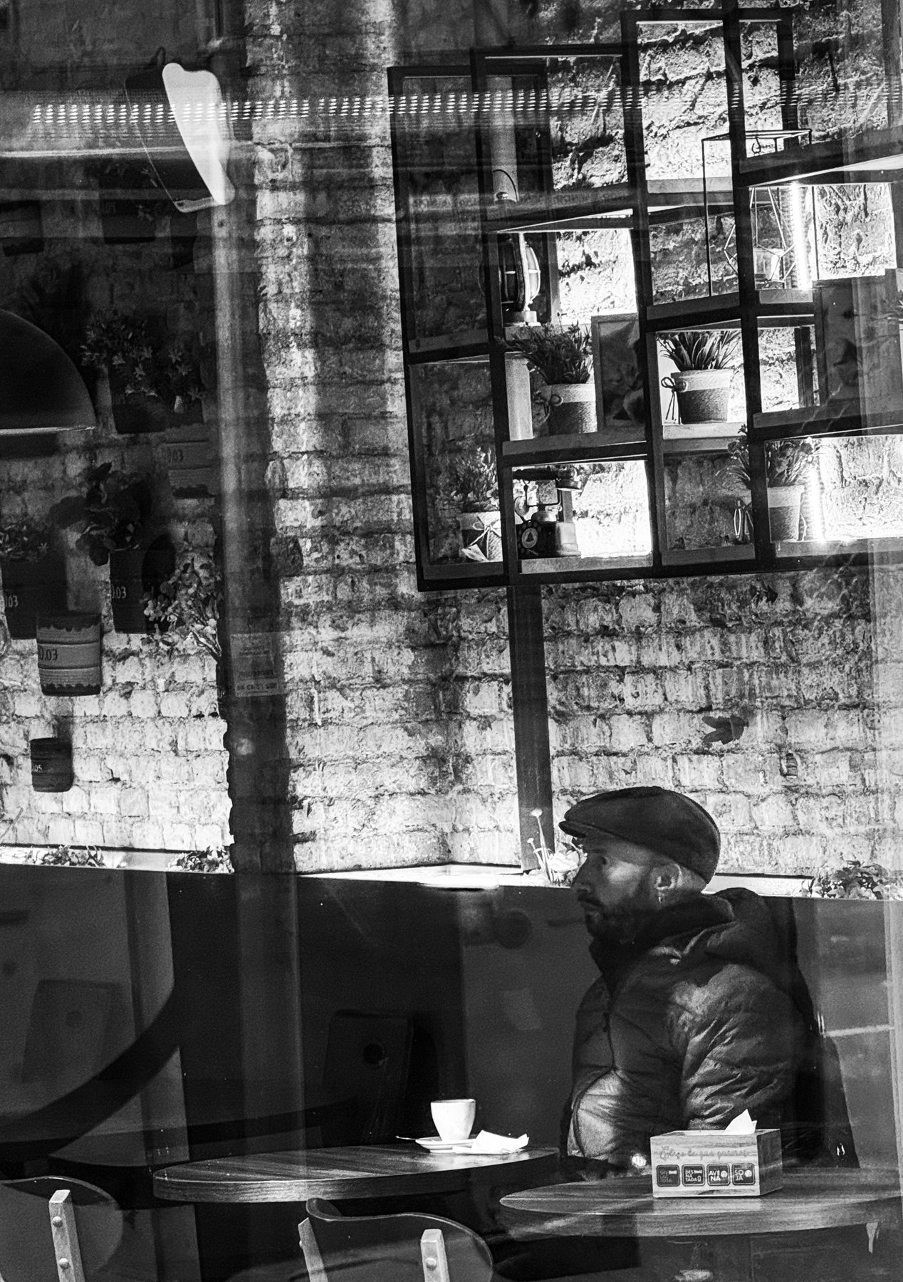 Media escena — hombre con gorra sentado con un café, visto a través del cristal de la cafetería con reflejos superpuestos, en blanco y negro