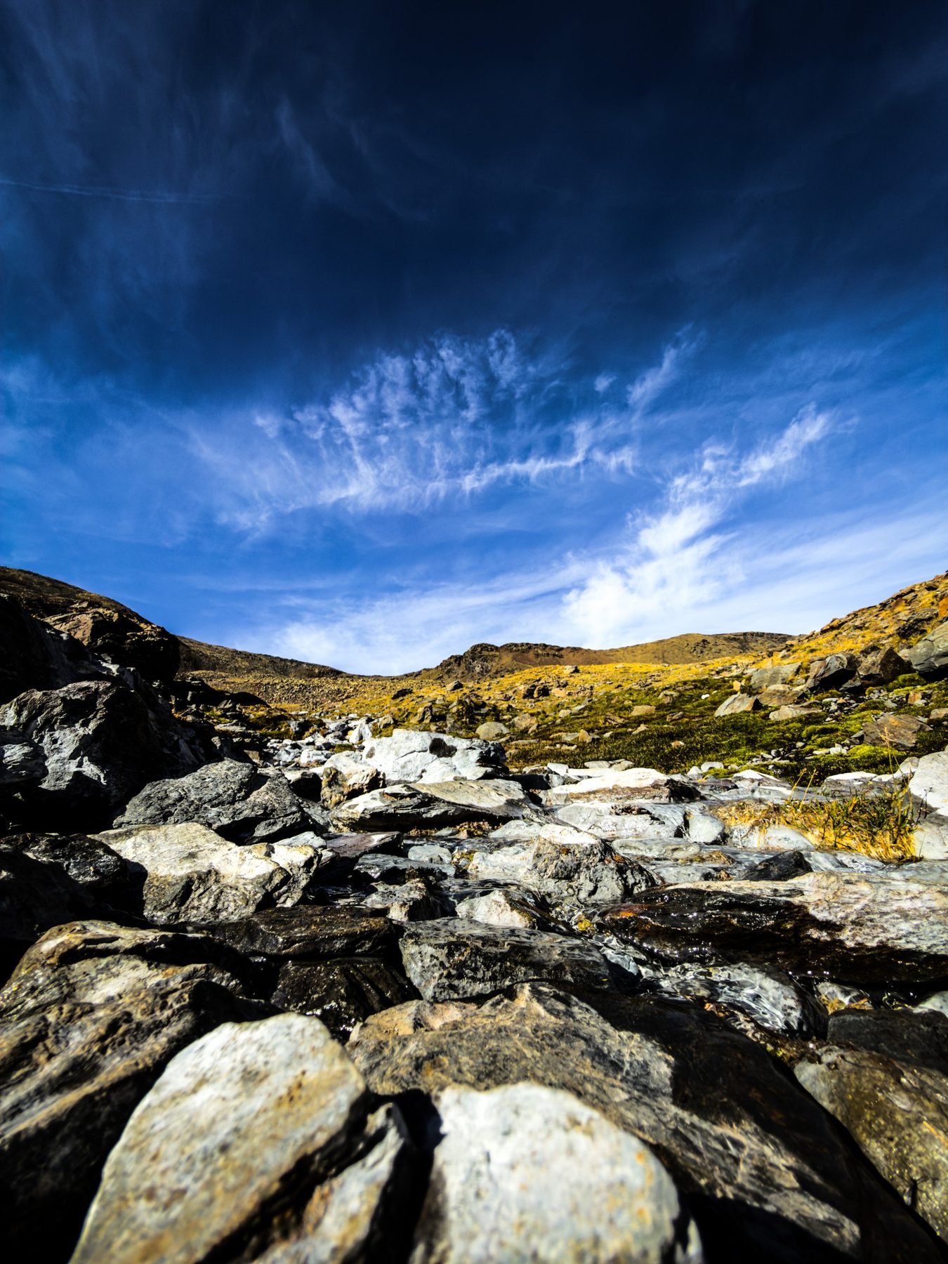 Cuesta arriba — lecho de piedras de un arroyo de alta montaña visto desde el suelo, con la ladera amarilla al fondo y un cielo azul intenso cruzado por nubes altas y finas, en color