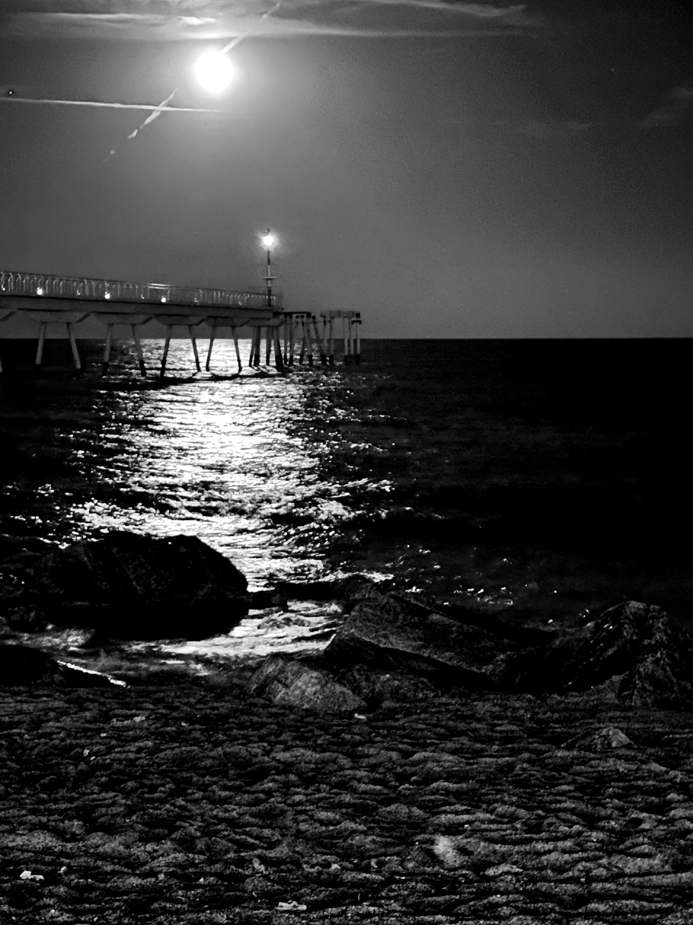 De noche — pasarela sobre el mar con la luna reflejada en una franja de luz sobre el agua, en blanco y negro