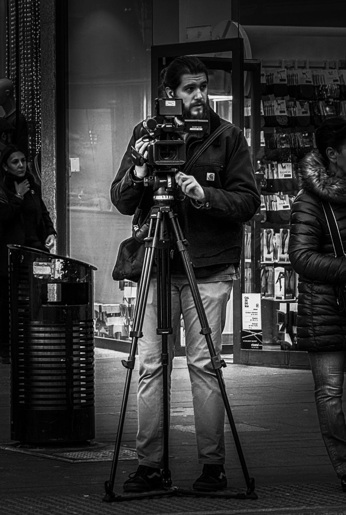 El que graba — cámara de vídeo sobre trípode manejada por un operador en plena acera comercial, en blanco y negro