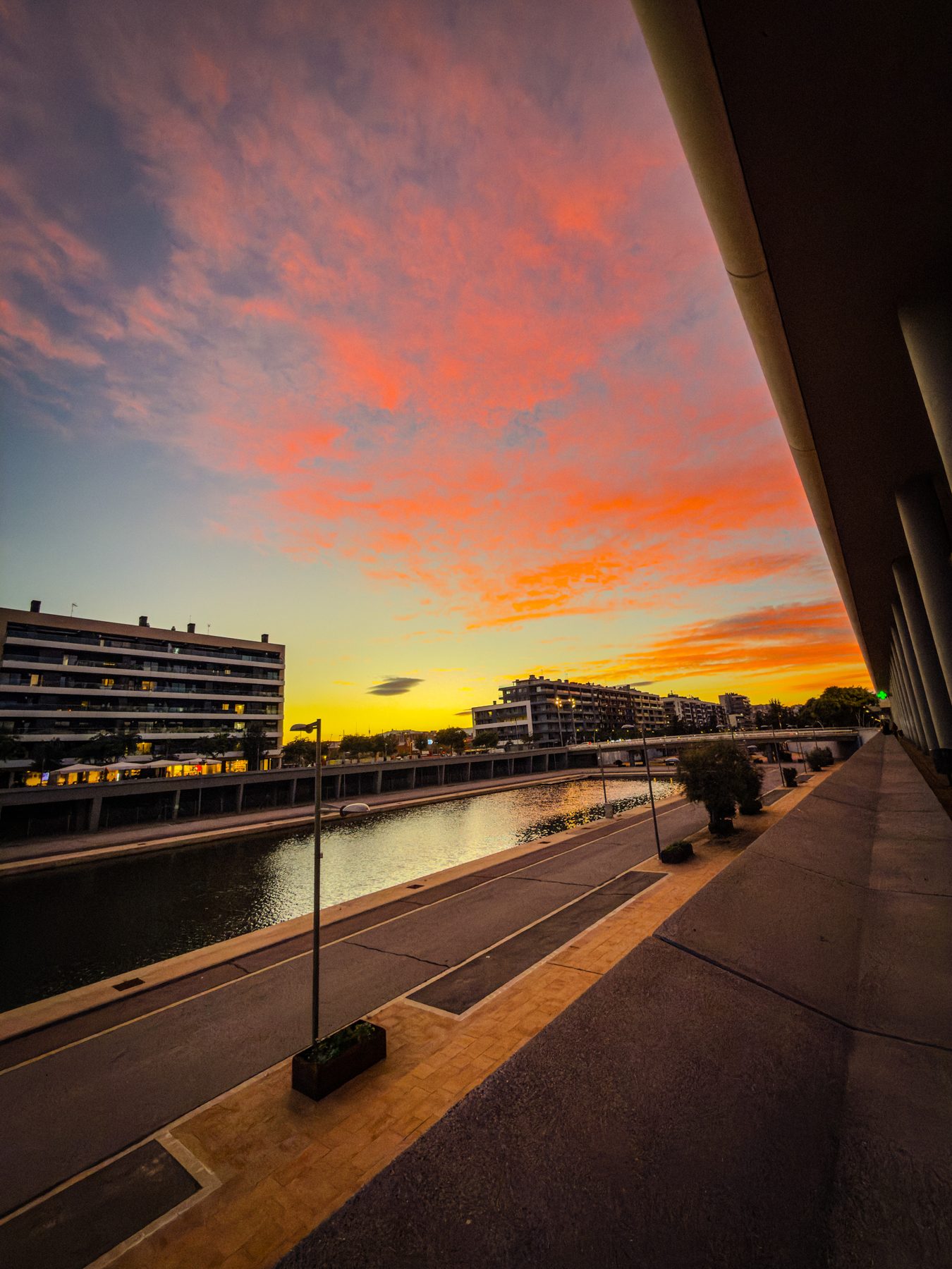 Por libre — atardecer con nubes naranjas y rosas sobre un canal, con una hilera de columnas en sombra a la derecha, farolas y bloques de pisos al fondo, en color