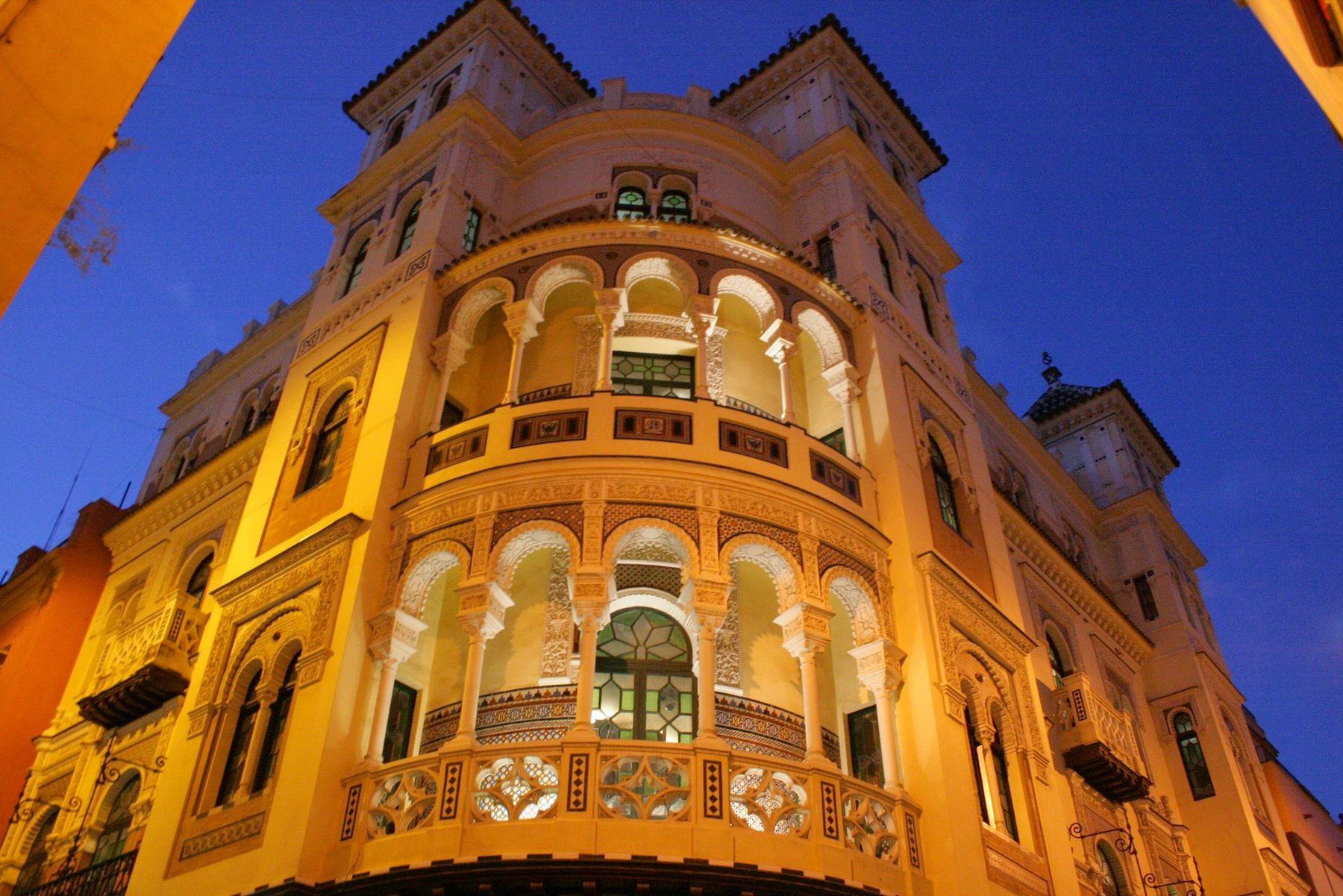 Esquina sevillana — edificio de arcos y balcones redondeados en una esquina de Sevilla, iluminado de amarillo y visto desde abajo contra el azul de la noche, en color