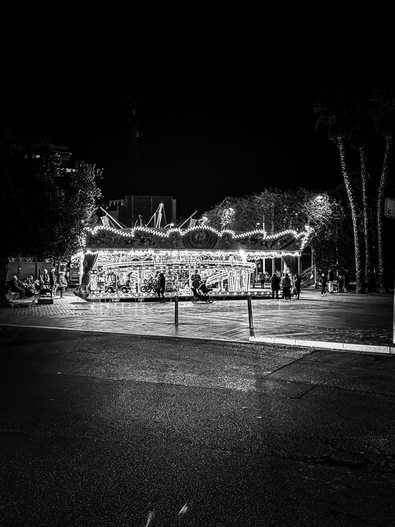La vuelta — carrusel iluminado en una plaza de noche, con el asfalto mojado en primer plano, en blanco y negro