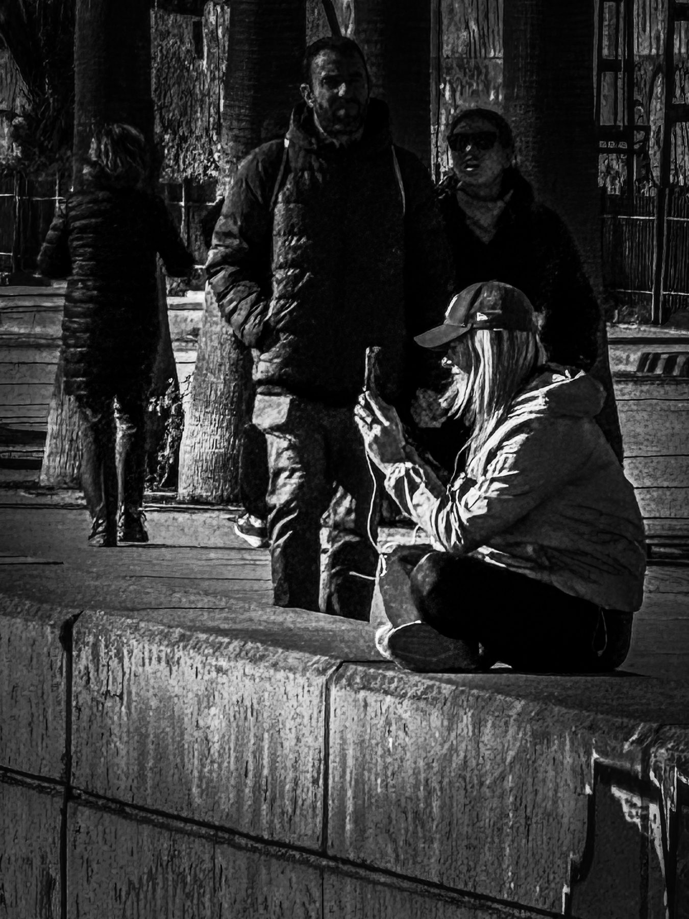 Al sol — persona con gorra sentada con las piernas cruzadas sobre un muro al sol mirando el móvil, con gente de pie detrás, en blanco y negro