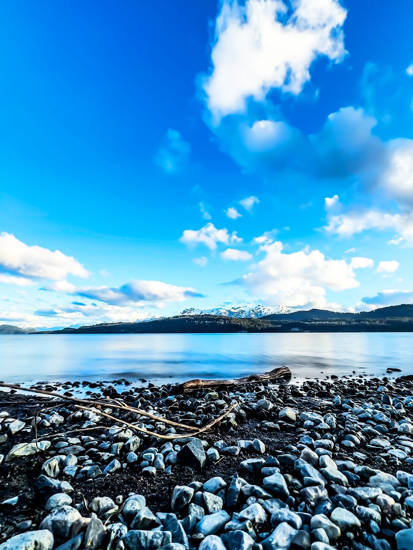 Orilla de piedra — playa de cantos rodados en primer plano, el lago Nahuel Huapi en calma y montañas nevadas bajo un cielo azul con nubes, en color