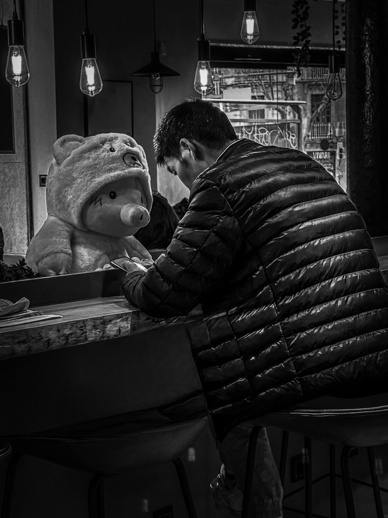 Compañía — hombre inclinado sobre el móvil en la barra de un bar, con un peluche grande sentado enfrente, en blanco y negro