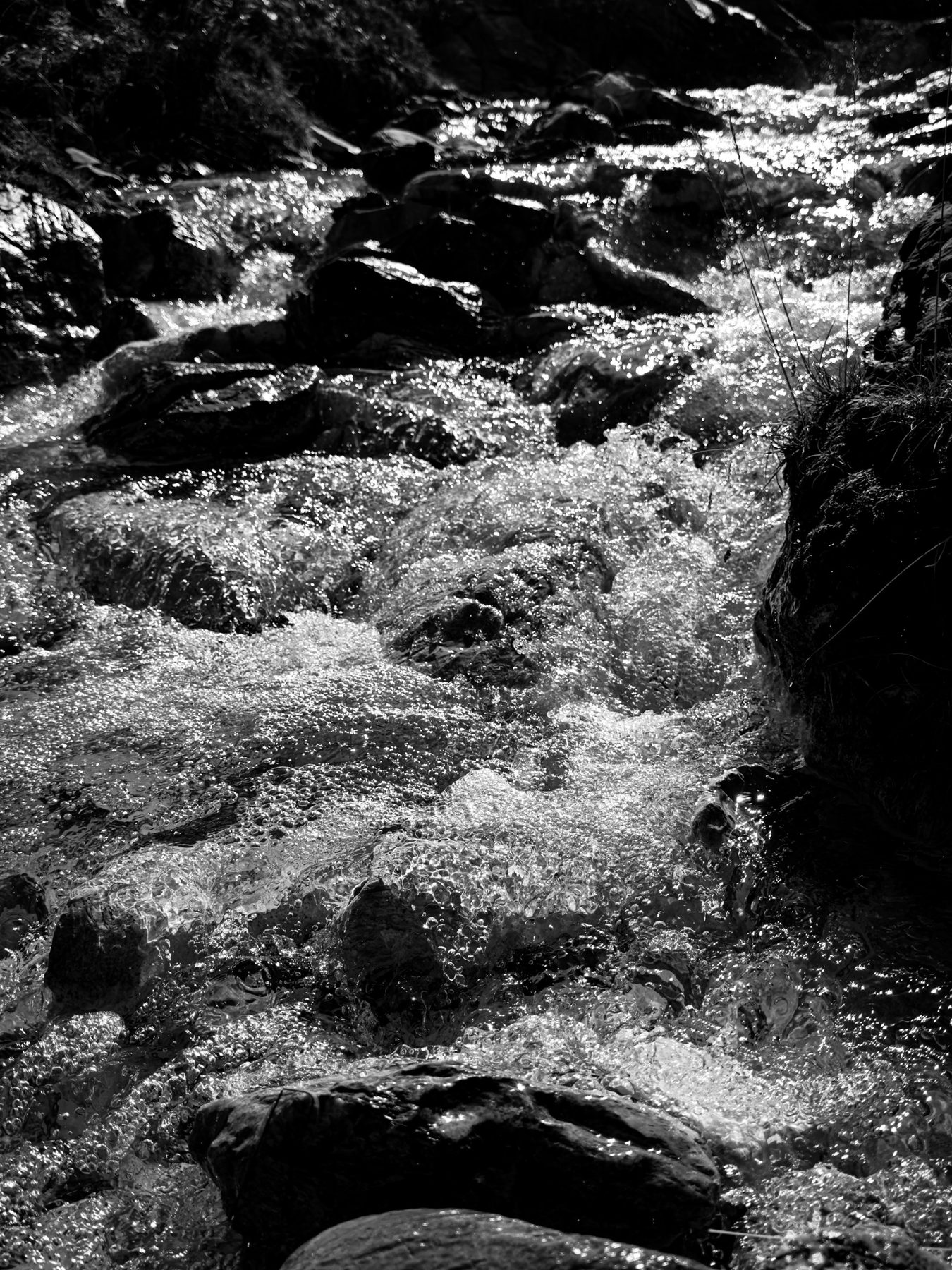 Por encima — arroyo de montaña bajando entre rocas, con el agua llena de burbujas y reflejos de sol, en blanco y negro de alto contraste