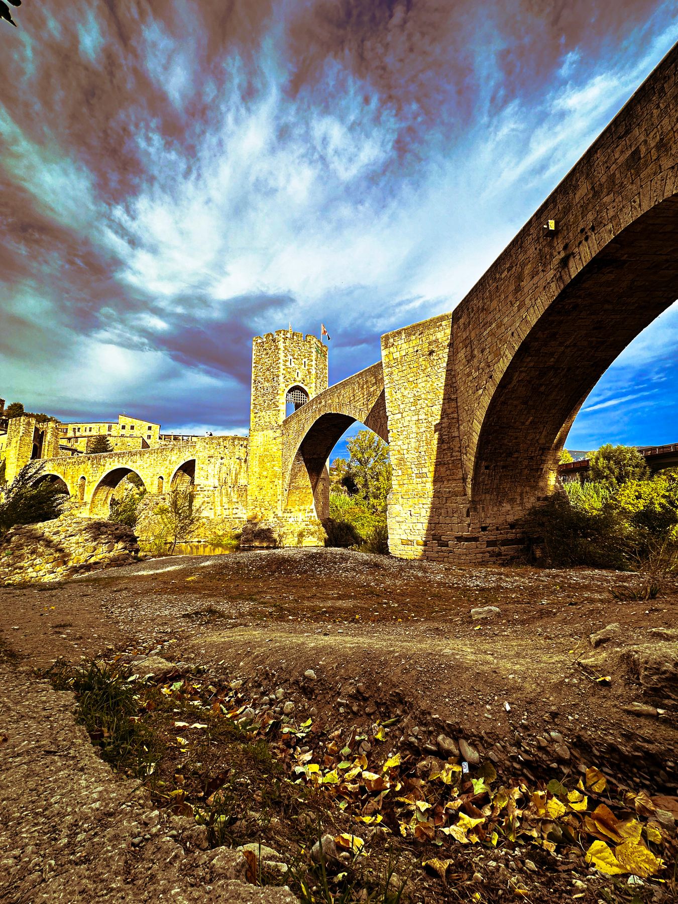 El paso — puente medieval de piedra sobre el cauce seco, con la torre almenada al fondo y hojas de otoño en primer plano
