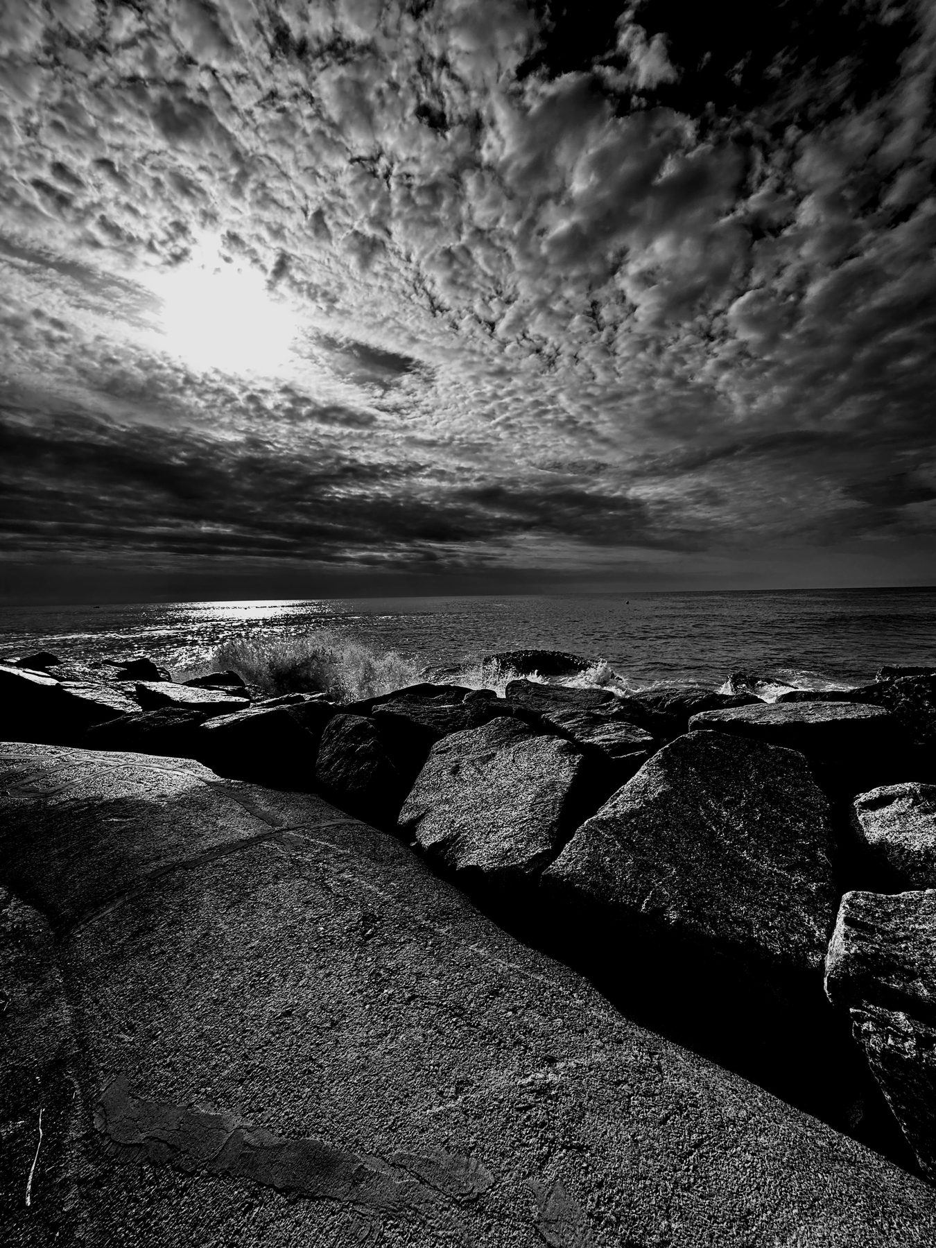 Las piedras — Bloques de piedra de un rompeolas y el sol tras las nubes, en blanco y negro