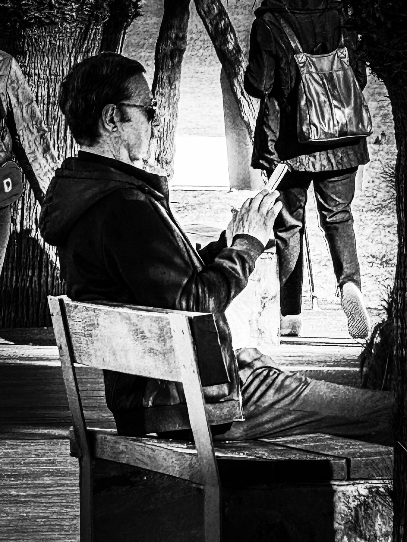 Sentado — hombre sentado en una silla de terraza mirando el móvil mientras la gente pasa, en blanco y negro