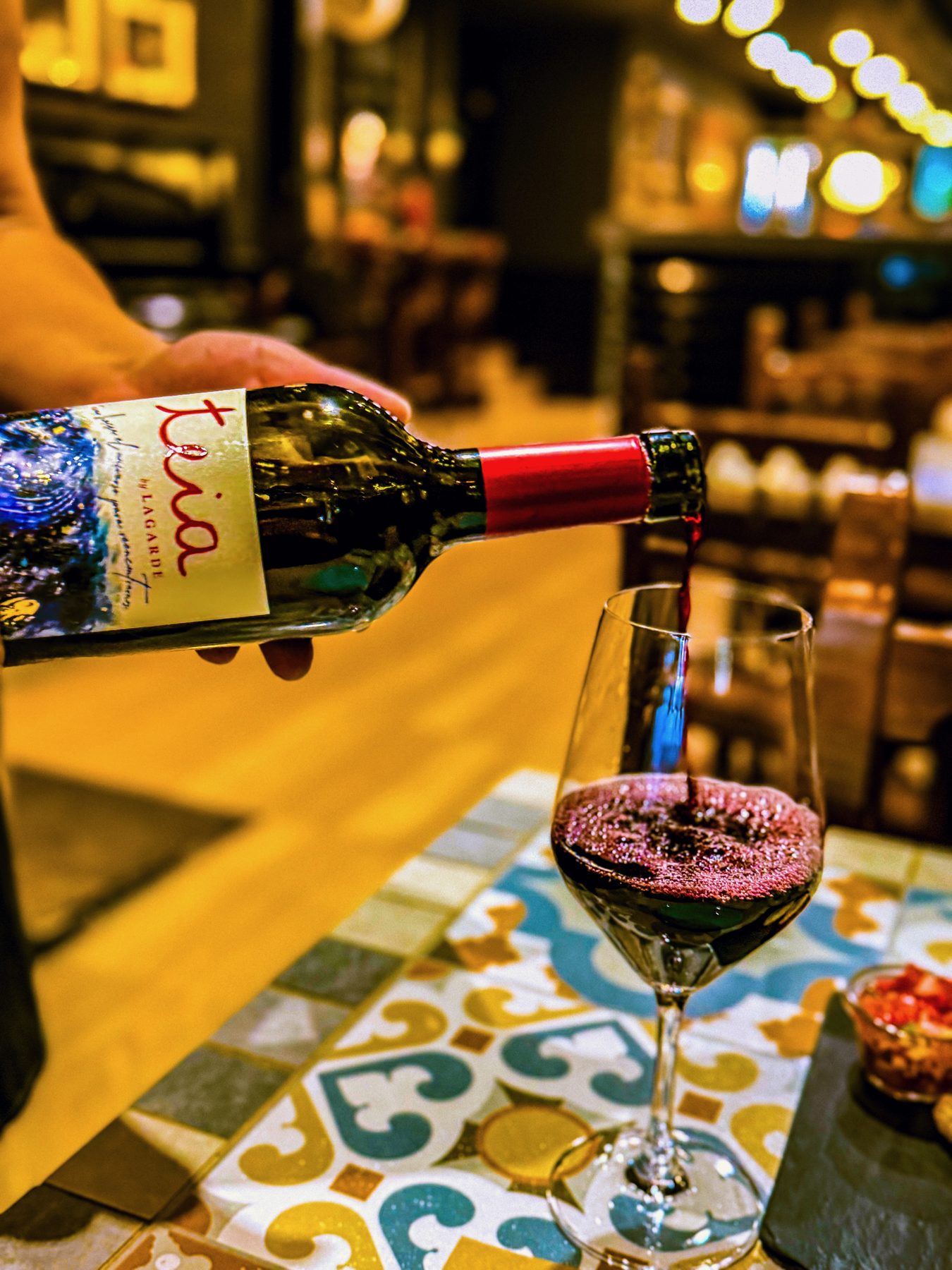 Servida — botella de tinto sirviéndose en una copa sobre una mesa de mosaico, en un bar con luces al fondo