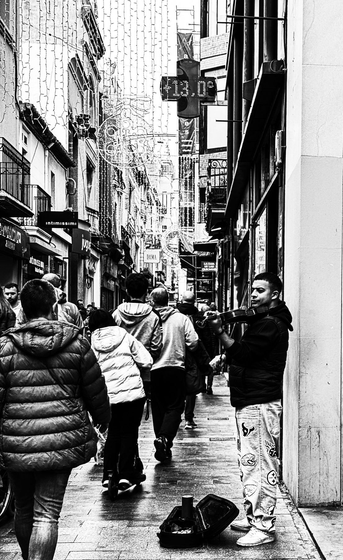 Contra la corriente — violinista tocando pegado a la pared en una calle comercial con luces de Navidad y gente de espaldas, en blanco y negro